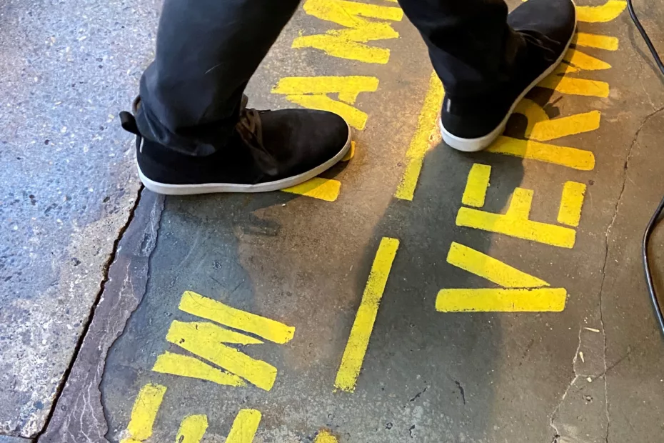 Feet stand on either side of a yeallow painted line in the Wehtstone Brewery taproom indicating the state line between VT & NH.